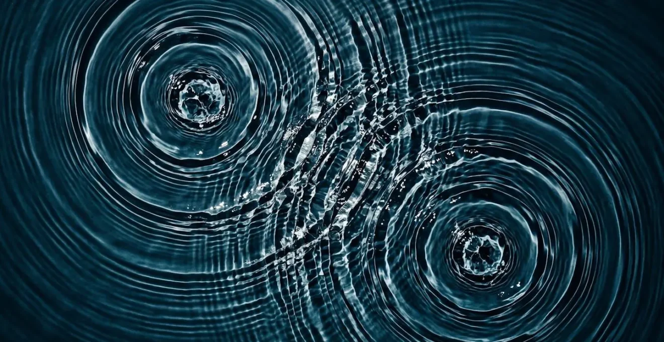 Vue aérienne d'ondes concentriques se propageant dans l'eau depuis deux points distincts, créant des interférences constructives et des vagues amplifiées à leur intersection